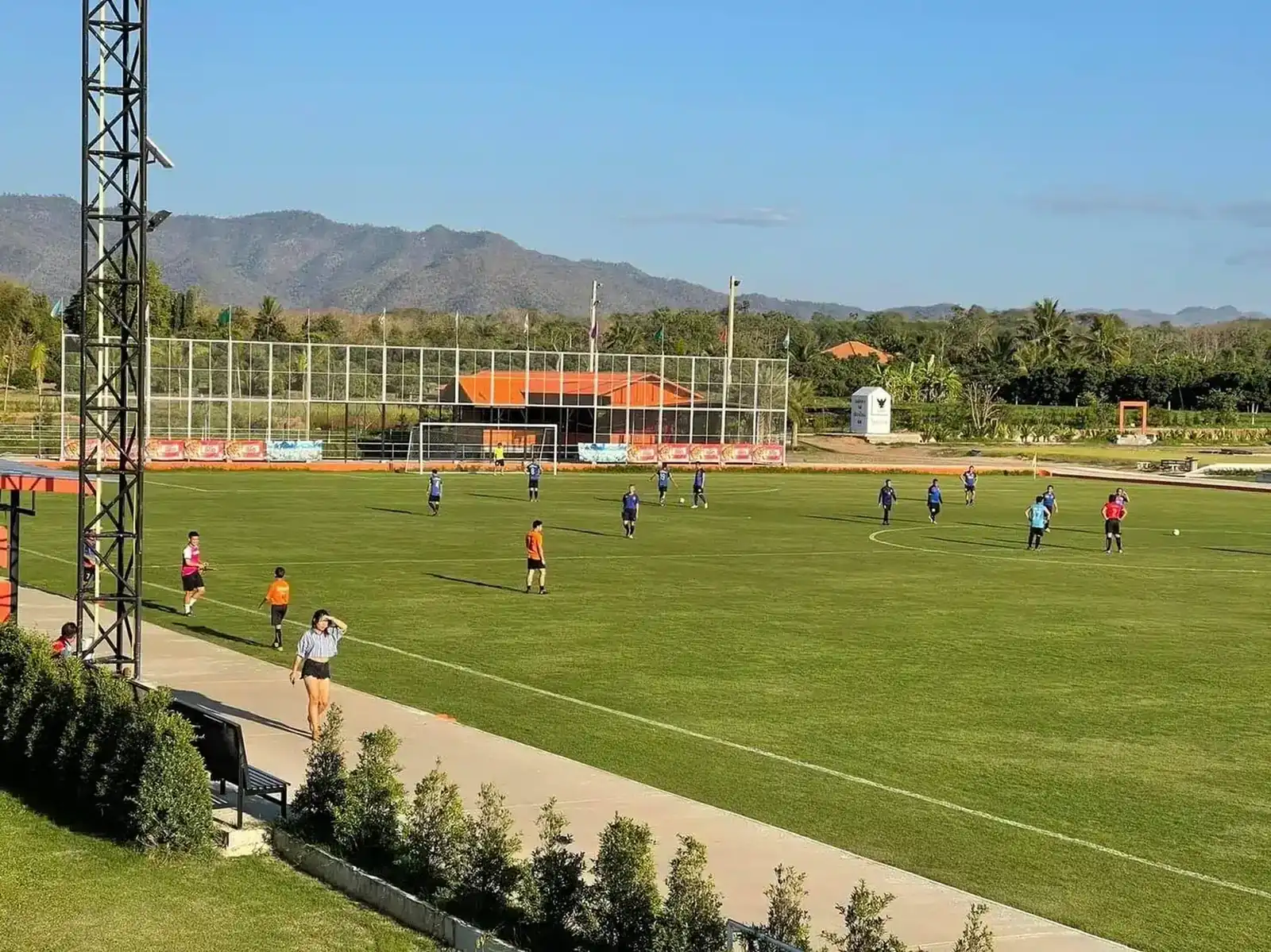 A squad training on the main pitch beside the clubhouse, a floodlight tower and mountains behind